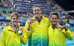 Image of 3 Australian Para-swimmers dressed in gold tracksuits, smiling at the camera and holding medals in their left handss.