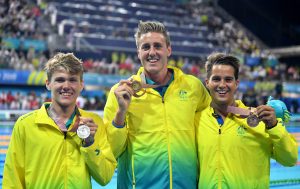 Image of 3 Australian Para-swimmers dressed in gold tracksuits, smiling at the camera and holding medals in their left handss.