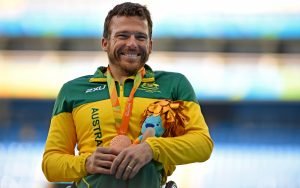 Male Paralympian Kurt Fearnley dressed in green and gold tracksuit representing Australia at the Rio 2016 Paralympic Games. Shown with a medal around his neck and a toy in his arms. He is smiling at the camera.