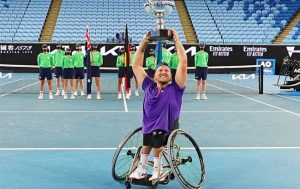 Australian wheelchair tennis player Dylan Alcott smiling at the camera and lifting a trophy above his head