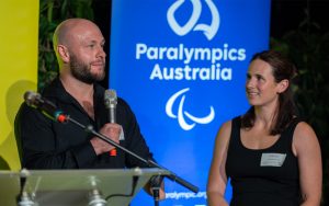 Image of male standing holding a microphone in his right hand speaking and a female standing to his left. They are both standing in front of a blue backdrop with the Paralympics Australia logo