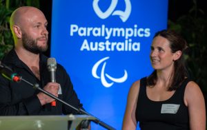 Image of male standing holding a microphone in his right hand speaking and a female standing to his left. They are both standing in front of a blue backdrop with the Paralympics Australia logo