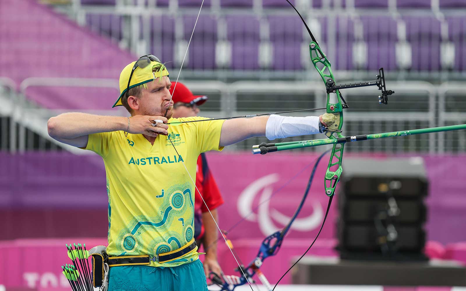 Australian Paralympian Taymon Kenton-Smith is wearing his green and gold Australian uniform. He is competing in an archery competition and aiming his arrow at the target ready to shoot.