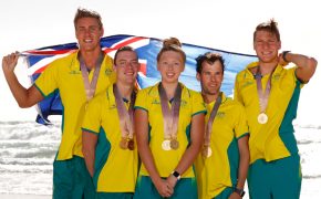 5 athletes wearing yellow polo shirts and green trackpants are standing in a group with the ocean as a backdrop smiling at the camera.